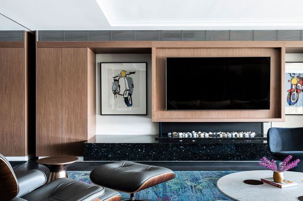 Contemporary living room with a wall-mounted television integrated into warm timber joinery, a glossy dark stone fireplace surround and hearth, black leather lounge chairs and ottoman, a patterned blue rug, and a round light-coloured coffee table. Artwork and decorative objects add restrained colour accents.