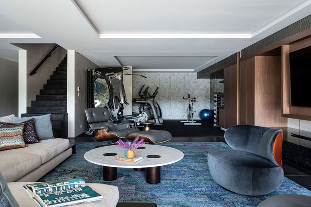 A sophisticated open-plan living room and home gym with a large sectional sofa, sculptural lounge chair, round coffee table, dark stone staircase, integrated timber television joinery, exercise equipment, and a textured feature wall. Recessed ceiling lighting and a richly patterned blue rug create a refined contemporary atmosphere.