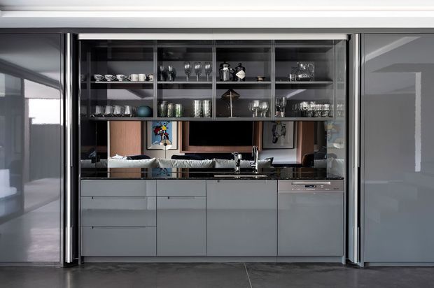 Contemporary compact kitchen and bar joinery viewed through large sliding glass doors. The space features smooth grey handleless cabinetry, a dark stone benchtop, integrated stainless-steel appliances, open glass-front shelving filled with glassware, and dark polished flooring. A living area is visible beyond the kitchen.