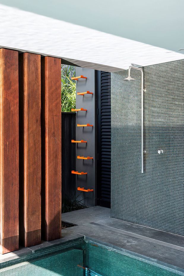 Modern outdoor pool area featuring a tiled outdoor shower, dark grey floor and wall mosaics, vertical timber screening, black louvred detailing, and bright orange wall-mounted accents. The pool edge and water occupy the foreground, creating a refined contemporary courtyard-like setting.