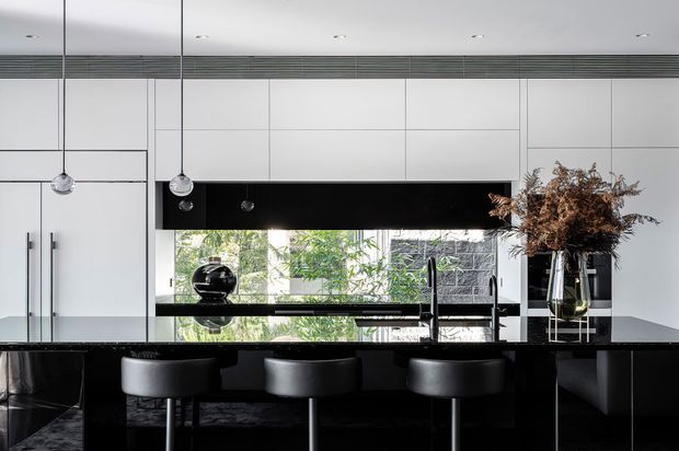 A sleek contemporary kitchen with a long black island and four bar stools, glossy dark cabinetry, white full-height cabinetry, integrated appliances, pendant lights, and a reflective splashback or window opening to greenery outside. The restrained black-and-white palette is softened by natural light and dried floral branches.