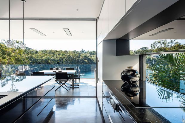 A sleek contemporary kitchen and dining area opening onto a pool terrace and lush wooded setting. The kitchen features dark glossy cabinetry, a polished dark stone island and benchtop, integrated appliances, reflective surfaces, and sculptural pendant lights. Full-height glazing and open edges create a strong connection to the outdoor pool and landscape.