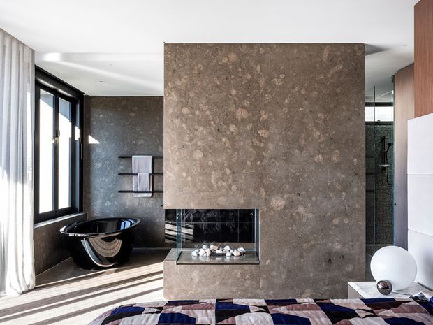 A contemporary bedroom with a freestanding black bathtub visible in an adjoining bathroom area. The room is organized around a large textured stone fireplace wall with an inset firebox, complemented by dark stone bathroom surfaces, black-framed glazing, timber joinery, and a patterned bedspread. Bright daylight enters through full-height windows and sheer curtains.