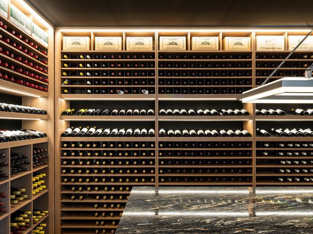 A sophisticated wine room or cellar lined with extensive floor-to-ceiling timber wine racks and illuminated bottle storage. Framed labels and wine displays sit above the shelving, while a large polished dark stone island or counter occupies the foreground. Warm integrated lighting creates an upscale, intimate atmosphere.