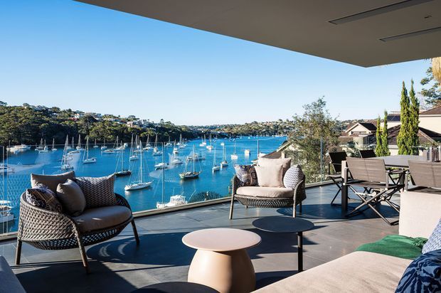 A spacious covered outdoor terrace overlooking a bright blue waterfront marina with numerous sailboats. The terrace features woven outdoor lounge chairs, a large upholstered sofa, small round coffee tables, and a dining or bar setting, with dark tiled or concrete-style flooring and a broad roof overhang. Residential buildings and landscaped vegetation are visible along the opposite shoreline.