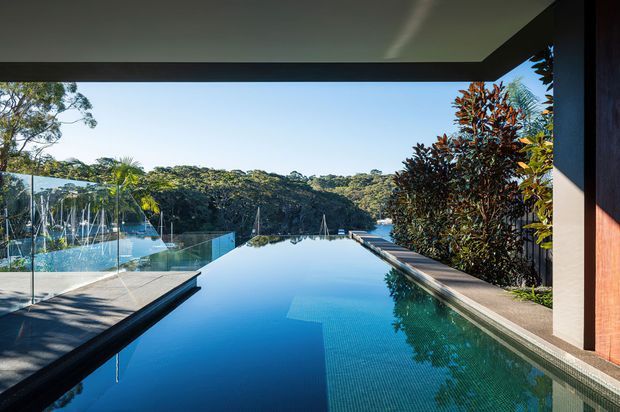 A modern architectural view from beneath a covered terrace, looking across a long rectangular infinity pool toward dense native bushland. The pool is bordered by dark paving and a narrow planted edge, with frameless glass pool fencing and contemporary timber-clad wall elements visible at the sides.