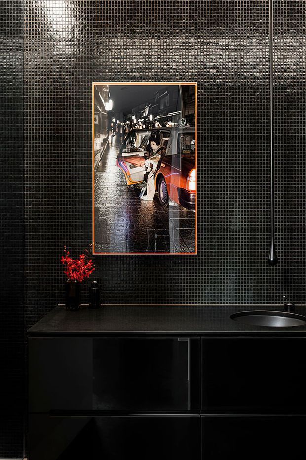 A dramatic, moody kitchen interior featuring glossy black small-format mosaic tile walls, sleek black handleless cabinetry, and a dark benchtop with an integrated or undermount sink. A framed urban night photograph adds a vivid orange accent, while a slim black pendant light hangs above the work surface.