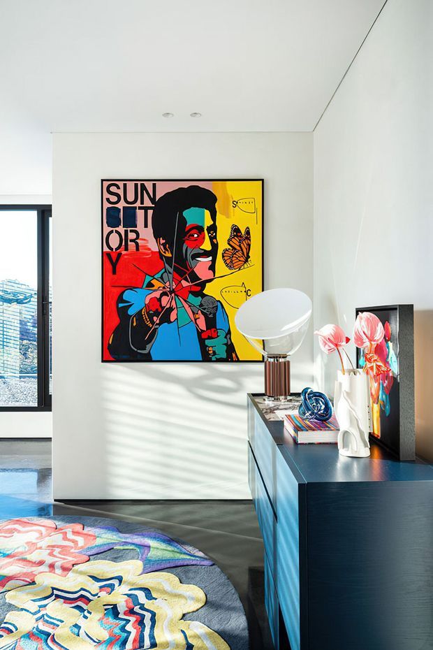 Bright contemporary bedroom corner with white walls and ceiling, a large colourful pop-art print, a dark blue built-in dresser, sculptural table lamp and decorative objects. Strong sunlight creates striped shadows across the wall and dark floor, while a vividly patterned bedspread occupies the foreground.