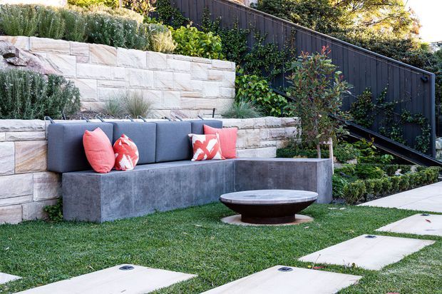 A landscaped outdoor entertaining area with a built-in concrete bench and dark upholstered cushions set against layered light stone retaining walls. Red patterned cushions add colour, while a low circular fire pit or coffee table sits on a manicured lawn beside large pale stepping slabs. Dense planting and a dark timber privacy fence complete the contemporary garden setting.