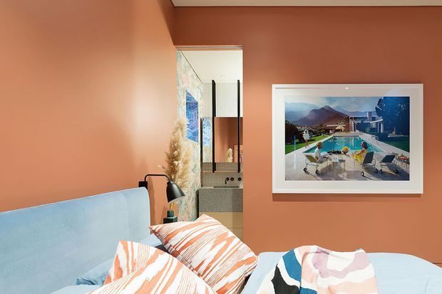 A warmly styled bedroom with terracotta-orange walls, a pale blue upholstered bed, layered abstract-patterned bedding, a black bedside lamp, and a framed landscape artwork. A mirrored or glazed opening reflects part of an adjacent vanity or bathroom area. The room has a colourful, contemporary retro-inspired feel.