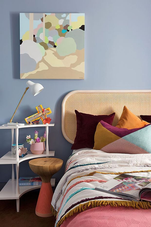 A colourful, eclectic bedroom vignette with a textured cane headboard, layered multicoloured cushions and a richly patterned bedspread. The room has a muted blue wall, abstract artwork, a white tiered bedside shelf with books and flowers, a sculptural two-tone timber side table, and a brass-and-white angled reading lamp.