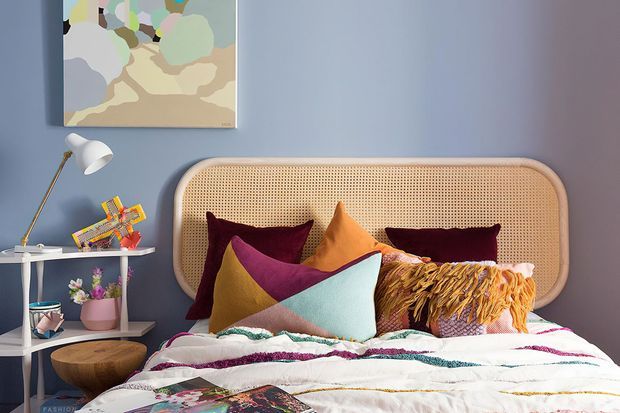 A colourful, eclectic bedroom with a rattan cane bedhead against a muted blue wall. The bed is layered with patterned white bedding, velvet and geometric cushions, and a mustard fringed throw. A slim side table with a white lamp and decorative objects sits beside the bed, while an abstract artwork adds colour above.