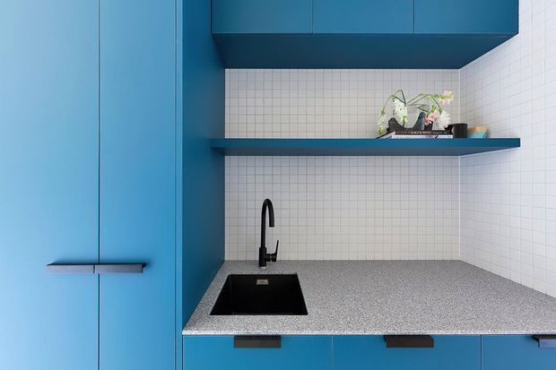 Compact contemporary kitchen or utility-style cabinetry with vivid blue flat-panel joinery, a speckled grey benchtop, black undermount sink and tap, and a white small-format tiled splashback. Open shelving and upper cabinets provide storage, with a small decorative plant on the shelf.