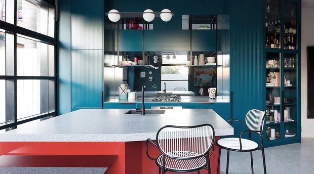 A bright contemporary kitchen with a large pale stone island, integrated seating, teal-blue fluted cabinetry and wall panels, open shelving, black-framed glazing, and a built-in bar cabinet. Red cabinetry and bold striped dining stools add colourful accents, while spherical pendant lights hang above the island.