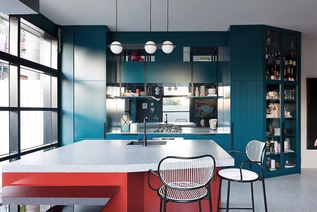 A contemporary kitchen with a large pale stone island, vivid red base cabinetry, deep teal fluted cabinetry, open shelving, a compact breakfast area, and full-height black-framed glazing. Globe pendant lights and ribbed metal chairs add graphic mid-century-inspired character.