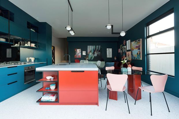 A vibrant open-plan kitchen and dining area with deep teal cabinetry and walls, a coral-red island with open shelving, pale stone benchtops, and a light terrazzo-style floor. The space includes a dining table with pink upholstered chairs, contemporary pendant lighting, large black-framed windows, and colourful artwork in the adjoining living area.