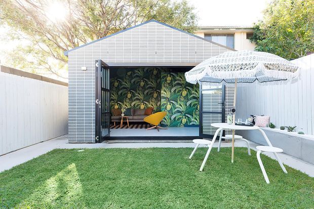 A bright backyard courtyard featuring a small contemporary gable-roofed garden studio clad in pale grey rectangular tiles. The studio has large black-framed glazed doors opening onto a compact interior with a botanical feature wall, timber bench seating and a mustard-yellow chair. In front is a manicured lawn, a white outdoor dining set and a fringed patterned patio umbrella, enclosed by white fencing.