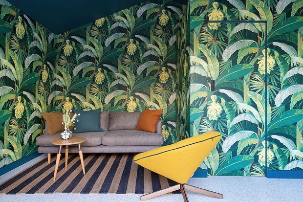 A compact, boldly styled living room with floor-to-ceiling tropical botanical wallpaper in deep green and yellow tones. A grey sofa with teal and burnt-orange cushions sits against the patterned walls, paired with a small round timber coffee table, striped area rug, and sculptural mustard-yellow lounge chair. The space has a dark ceiling and a playful, high-contrast interior scheme.