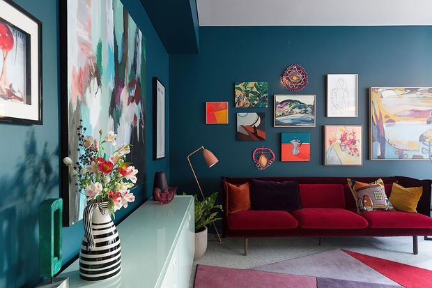 Eclectic living room with deep teal walls, a red velvet sofa, colourful abstract and figurative artwork arranged as a gallery wall, a white built-in sideboard, patterned rug, sculptural ceramic vase, and warm decorative lighting. The room has a bold, artistic and richly layered interior.