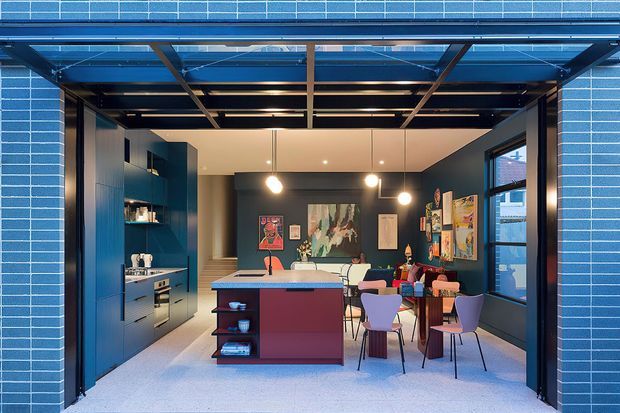 A contemporary kitchen and dining area viewed through fully opened glazed garage-style doors. The space features dark blue built-in cabinetry, a central island with a deep red base and pale benchtop, pendant lighting, colorful artwork, a dining table with mismatched chairs, and a blue glazed brick exterior frame. The large opening creates a strong indoor-outdoor connection.