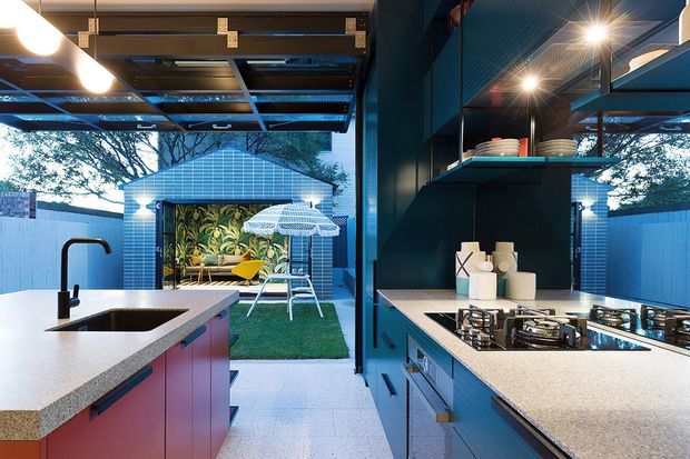 A compact contemporary kitchen with a galley-style layout, dark blue-green cabinetry and walls, pale stone-look benchtops, an undermount sink, gas cooktop, and open shelving. Full-height glazed doors open to a small landscaped courtyard with a patterned outdoor pavilion, seating, and umbrella. Exposed overhead structure and strong indoor-outdoor connection are prominent.