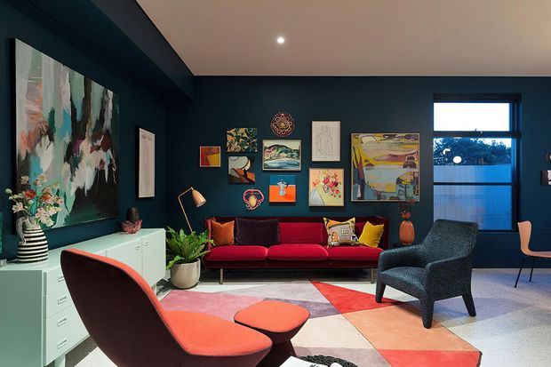 A richly layered, moody living room with deep navy walls, a gallery wall of colourful artwork, a red and burgundy sofa, patterned rug, sculptural lounge chair and ottoman, and a large abstract artwork. A window brings in natural light, while a slim floor lamp and recessed ceiling light illuminate the room.