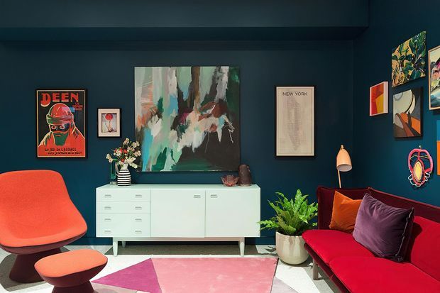 Eclectic, richly coloured living room with deep navy walls, a low pale built-in sideboard, abstract artwork, framed posters, a bright red sofa with dark cushions, and an orange mid-century-style lounge chair and ottoman. A colourful geometric rug, indoor plants, sculptural lamp, and layered wall art create a bold, curated atmosphere.