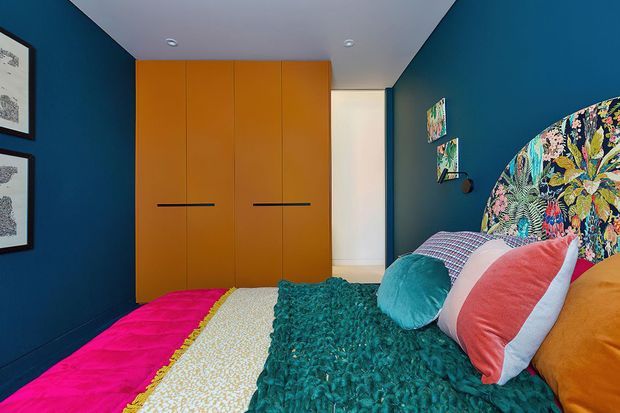 A compact, vividly styled bedroom with deep blue walls, a large floral upholstered headboard, and a bed layered with bright pink, cream, teal, coral, turquoise, and orange textiles. Floor-to-ceiling orange built-in wardrobe cabinetry occupies the rear wall, with framed artwork and small wall-mounted lights adding detail. The room has a standard-height white ceiling and a bold, playful, eclectic character.