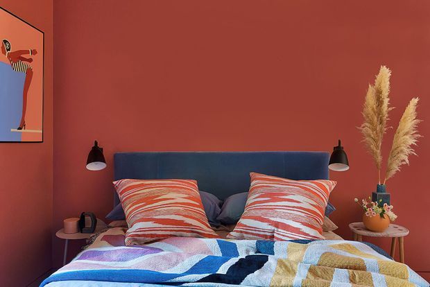 A bold, colourful bedroom with terracotta-red walls, a deep blue upholstered bedhead, patterned red-and-white cushions, and a vibrant geometric bedcover. Black wall-mounted bedside lamps flank the bed, while a small round side table holds decorative objects and tall dried pampas stems. A colourful framed artwork is partially visible on the left wall.