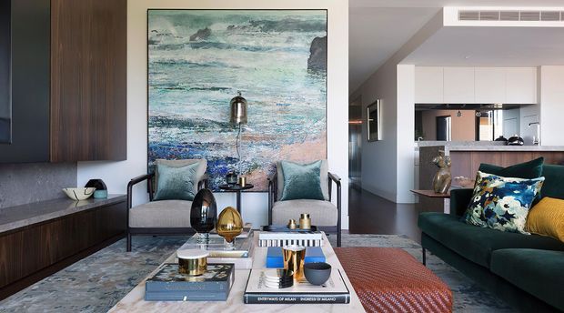 A refined open-plan living room with two armchairs facing a low coffee table and a deep green sofa in the foreground. The room features dark timber built-in joinery, a grey stone-like media wall and cabinetry, a large coastal abstract artwork, layered rugs, decorative cushions, sculptural accessories, and a kitchen visible in the background.