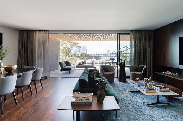 A refined contemporary open-plan living and dining room with expansive floor-to-ceiling glazing overlooking an urban waterfront or cityscape. The room features dark timber flooring, a deep green sectional sofa, sculptural lounge chairs, a patterned blue-grey rug, a sleek low coffee table, and a dark built-in media wall. Full-height curtains frame the glazing, while the dining area sits to the left with a modern table and upholstered chairs.