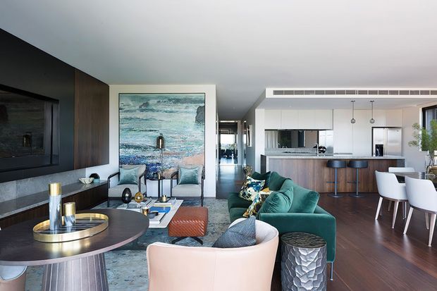Contemporary open-plan living and dining area flowing into a bright kitchen. The room features dark timber flooring, a charcoal feature wall with built-in television joinery, a teal sectional sofa, sculptural occasional tables, a round dining table, and a white kitchen with a timber island. Large abstract coastal artwork and extensive glazing add colour and light.