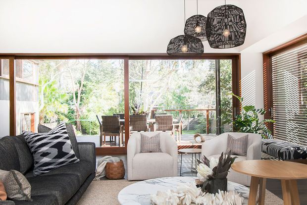 Bright, warmly styled living room with expansive timber-framed glazing opening toward a leafy outdoor setting. The room features a dark textured sofa, two light upholstered armchairs, a round marble coffee table, timber side and dining tables, indoor plants, woven accents, timber venetian blinds, and three sculptural black woven pendant lights.
