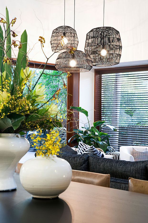 A warmly styled living and dining area with a dark wood-framed window, horizontal blinds, a charcoal sofa, leather dining chairs, and a timber dining table. Three sculptural woven pendant lights hang overhead, while large ceramic vases and lush botanical arrangements add organic detail. The space is bright, layered, and contemporary with strong natural materials and textural accents.