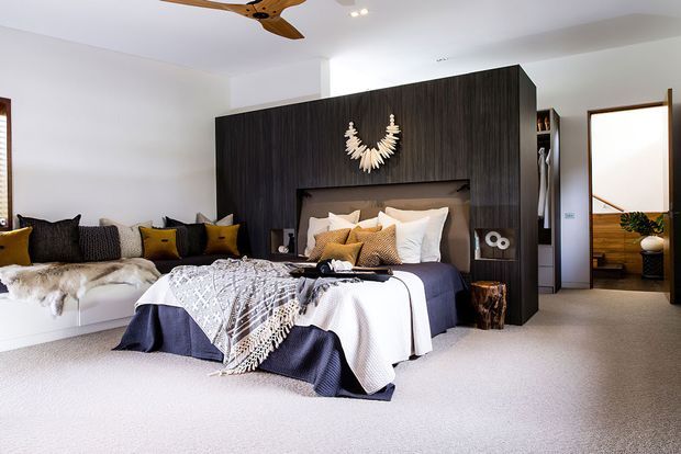 A bright, spacious contemporary bedroom with a large dark timber feature wall and integrated headboard cabinetry. The bed is layered with white, navy, mustard and patterned textiles, while a built-in window seat with cushions sits along the left wall. A timber ceiling fan, recessed lighting, carpeted flooring and an open doorway to an adjacent space complete the room.