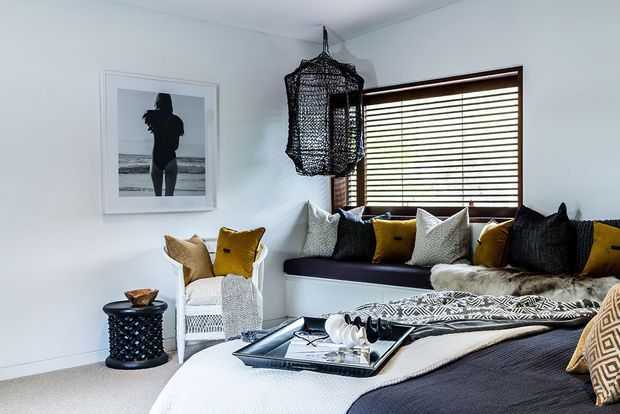 A cozy, stylish bedroom with a built-in window seat beneath horizontal timber blinds, layered black, white, grey and mustard cushions, a woven pendant light, patterned textiles, and a large upholstered bed. The room has white walls, light carpet, and a relaxed coastal-inspired monochrome palette with warm accents.