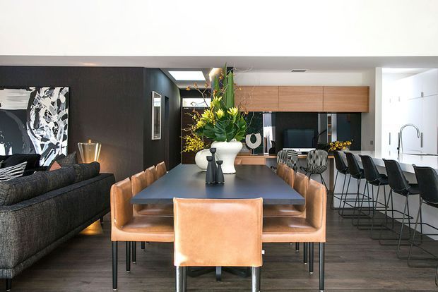 Open-plan contemporary dining and kitchen interior with a long dark dining table surrounded by tan leather chairs, a dark sectional sofa at left, black feature walls, timber overhead cabinetry, and a bright white kitchen with a large island and bar stools. Indoor planting and artwork add decorative contrast.