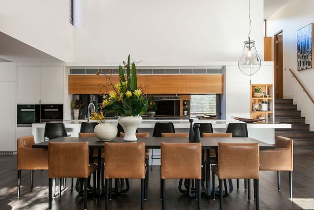 Open-plan dining and kitchen interior with a large timber dining table surrounded by brown leather chairs. A contemporary kitchen sits behind it with white cabinetry, a timber feature joinery band, dark benchtops, integrated appliances, pendant lighting, and a staircase leading upward at the right. The room has high ceilings and a refined neutral palette with warm timber and leather accents.