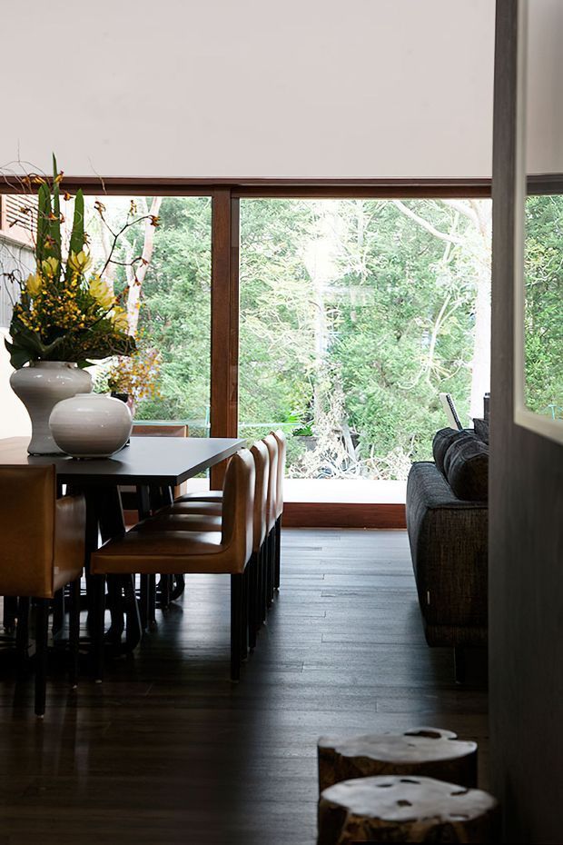 A refined open-plan dining and living area with a dark timber dining table, upholstered dining chairs, sculptural ceramic vases, and a dark sofa. Full-height timber-framed glazing overlooks dense greenery, while dark timber flooring and a neutral ceiling create a warm, nature-connected interior.