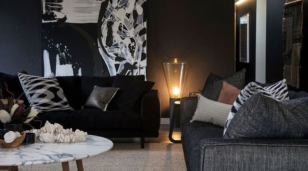 A dark, refined living room with charcoal sofas, monochrome patterned cushions, a round white marble coffee table, sculptural decor, and a warm floor lamp. Black-and-white abstract artwork and deep-toned walls create a dramatic, sophisticated interior.