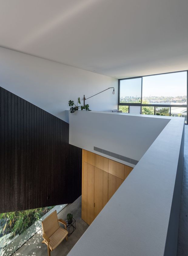 A bright contemporary stairway and landing overlooking a double-height void. A dark vertical timber-paneled stair wall contrasts with white plastered balustrades and walls, while warm timber cabinetry sits below. Large black-framed windows provide views over a leafy urban landscape, and a small seating area with plants is visible at the lower level.