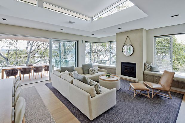 Bright, spacious open-plan living and dining area with floor-to-ceiling glazing, a large skylight, pale timber flooring, a neutral sectional sofa, built-in window seating, and a central fireplace feature wall. The dining area is visible to the left, surrounded by views of trees.