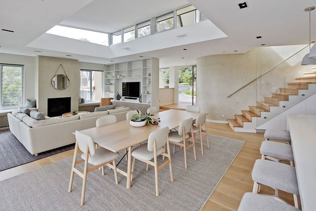 Bright open-plan residential interior combining a dining area, living room and stairway. The space features a large skylight and clerestory glazing, white built-in shelving, a fireplace with a circular mirror, pale upholstered furniture, a light timber dining table and chairs, and a sculptural open timber staircase against a softly textured plaster wall.