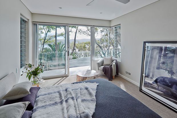 Bright contemporary bedroom with a large upholstered bed, layered blue-grey bedding, a textured throw, and a small bedside table. Full-height sliding glass doors open to a balcony with leafy outdoor views, while corner glazing brings in abundant natural light. A lounge chair and framed artwork complete the calm, refined space.