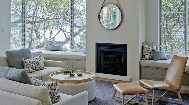 A bright, refined living room centred around a contemporary fireplace, with expansive windows overlooking trees, built-in window seating, two pale upholstered armchairs, a round coffee table, patterned cushions, and a tan leather lounge chair with matching ottoman. The palette is soft and neutral with light grey textiles, white walls, natural timber accents, and a textured rug.