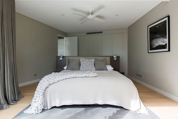 A bright, contemporary bedroom with a neatly made bed, layered grey and white bedding, full-height grey curtains, built-in pale cabinetry behind the bed, timber flooring, recessed ceiling lights, and a white ceiling fan. A monochrome framed artwork provides a restrained wall accent.
