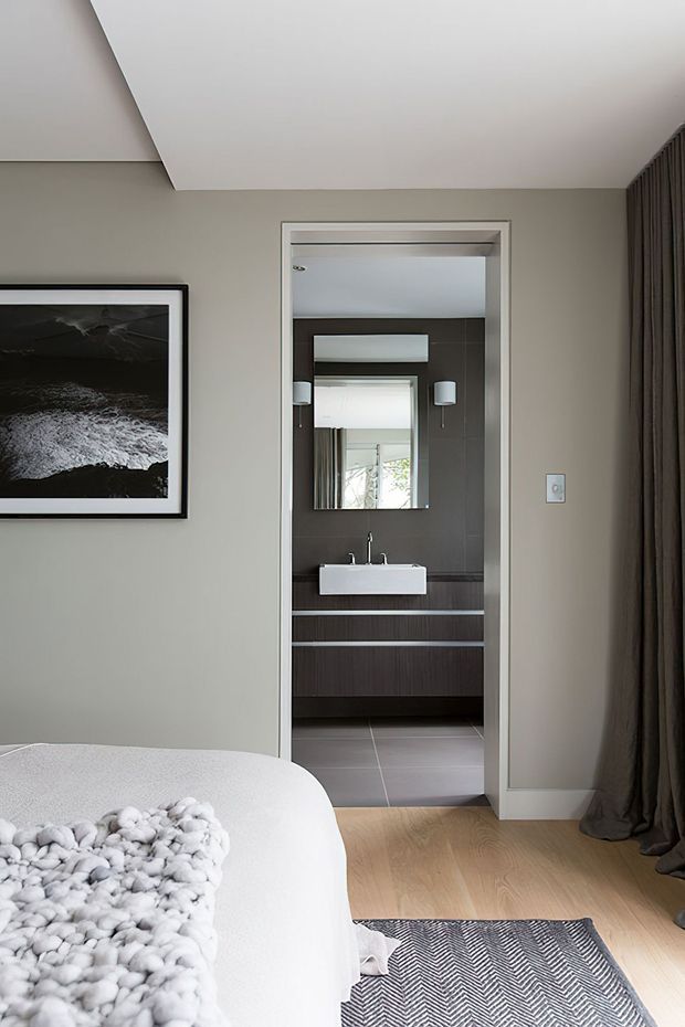 A contemporary bedroom viewed toward an open bathroom doorway. The bedroom has soft grey walls, a sloped white ceiling, pale timber flooring, a white upholstered bed with a chunky knitted throw, dark curtains, framed monochrome artwork, and a patterned rug. The adjoining bathroom features dark wood-look vanity cabinetry, a white wall-mounted basin, large grey tiles, a wide mirror, and modern wall sconces.