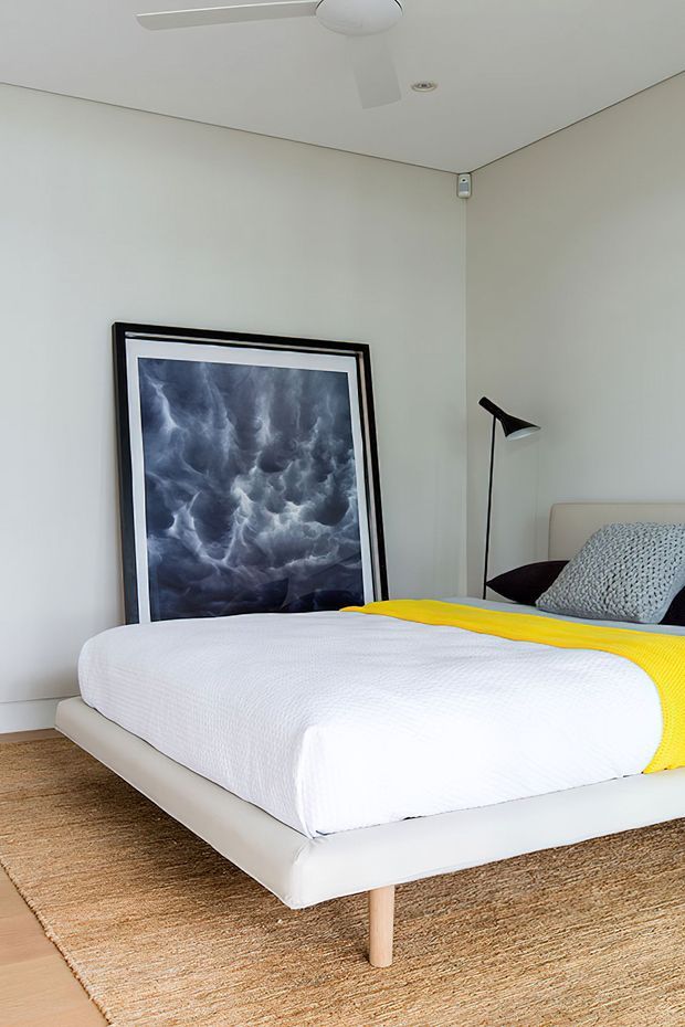 Bright, contemporary bedroom with a low upholstered platform bed dressed in white bedding and a vivid yellow throw. The room has pale neutral walls, a woven natural-fibre rug, a black minimalist floor lamp, framed monochrome artwork leaning against the wall, and a ceiling fan.
