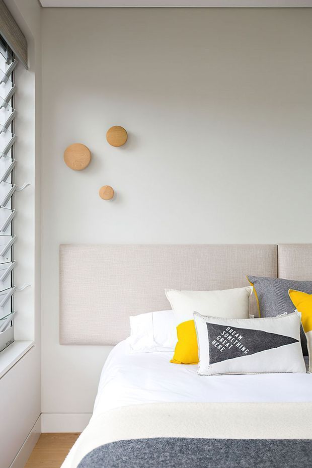 Bright, compact contemporary bedroom with a pale upholstered headboard, crisp white bedding, charcoal and yellow accent cushions, and simple circular timber wall decor. A louvred window provides natural light, while light timber flooring and soft neutral walls create an airy, clean atmosphere.