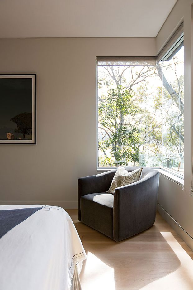 A bright, understated bedroom corner with a dark upholstered armchair positioned beside large corner windows. The room has warm beige walls, light timber flooring, a white-covered bed in the foreground, and a framed artwork on the wall. Abundant natural light and leafy trees are visible outside the glazing.
